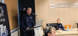 policjant w mundurze stoi przed mównicą, obok ekran, policjantka za stołem.