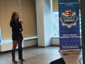 młoda kobieta w sukience trzyma w ręku mikrofon, w tle napis komenda powiatowa policji w Opocznie.