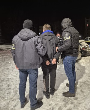 dwóch mężczyzn z napisem na rękawie policja trzyma mężczyznę w kajdankach na rękach trzymanych z tyłu.Stoją przed samochodami na śniegu.