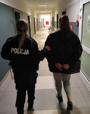 policjantka prowadzi kobietę, która ma założone kajdanki na rękach trzymanych z tyłu.