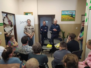 policjanci z stoją z mężczyzną przed siedzącymi na krzesłach osobami.