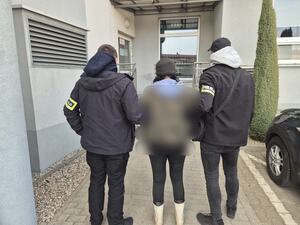 dwóch mężczyzn w ciemnym ubraniu z napisem na rękach policja, prowadzi kobietę, w czapce, przed budynkiem.