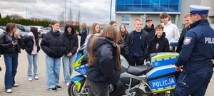 młodzież z policjantem znajdują się na parkingu, jedna dziewczyna siedzi na motocyku służbowym.