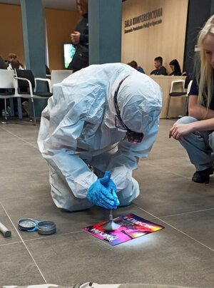 osoba ubrana w biały kombinezon i rękawiczki trzyma w ręku pędzelek, na podłodze znajduje się gazeta, w tle widoczne osoby, napis sala konferencyjna.