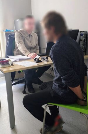 mężczyzna z kajdankami na rękach siedzi na krześle, za biurkiem siedzi mężczyzna w jasnej bluzie, na biurku dokumenty, przedmioty biurowe.