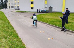 dziecko jedzie rowerem, obok policjanci, budynek, osoby stoją pod budynkiem.