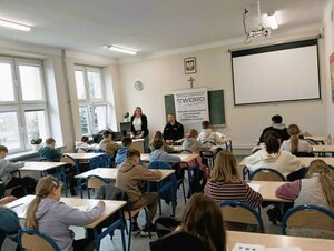 dzieci siedzą w szkolnych ławkach w klasie, kobieta i policjant stoją przed nimi.