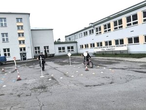 dziecko na rowerze pokonuje tor przeszkód, w tle budynek.