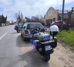 dwa policyjne motocykle. dwa samochody, droga asfaltowa, przy drodze sklep., w tle inne budynki.