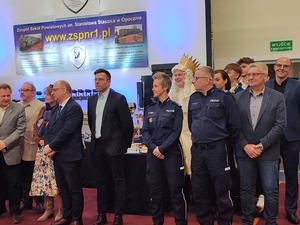 grupa ludzi, wśród nich policjant i policjantka, za nimi telebim.