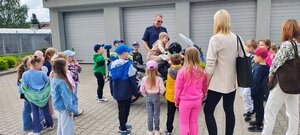 dzieci wraz z kobietami i policjantem znajdują się przed budynkiem, na motocyklu siedzi chłopiec.