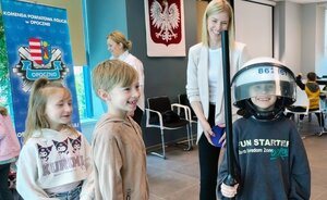 dziecko ubrane w hełm policyjny, w ręku trzyma pałkę , za nim kobieta.