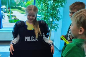 dziewczynka w kamizelce z napisem policja, obok chłopcy, wszyscy znajdują się w budynku komendy policji.