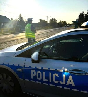 radiowóz policyjny stoi przy drodze, za nim policjant w odblaskowej kamizelce.