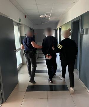 mężczyzna w kajdankach prowadzony jest korytarzem przez policjanta i policjantkę.