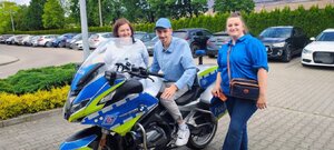 chłopiec siedzi na motocyklu policyjnym, obok stoją dwie kobiety, za nimi rząd samochodów.