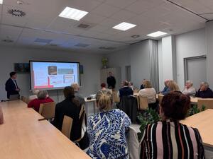 grupa ludzi siedzi za stolami, przed nimi widoczny ekran z wyświetlanymi napisami.