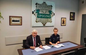 policjantka i mężczyzna w garniturze podpisują dokumenty, na ścianie widoczne logo policji.