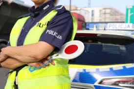 policjant w kamizelce z tarczką, za nim radiowóz.