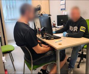 mężczyzna z napisem na rękawie policja siedzi przy biurku, na przeciwko siedzi mężczyzna w koszulce z krótkim rękawem.