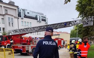 policjant stoi na tle budynku i samochodów strażackich, obok ratownicy.