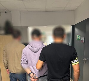 trzech mężczyzn, jeden w kajdankach, jeden z opaska na ręce z napisem policja.