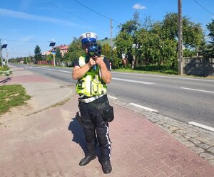 policjant w odblaskowej kamizelce stoi przy drodze, mierzy prędkość.