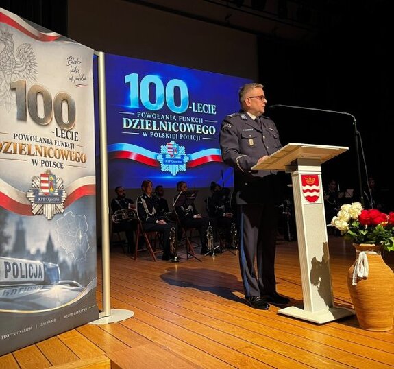 policjant stoi przy mównicy, za nim napisy o jubileuszu dzielnicowych.
