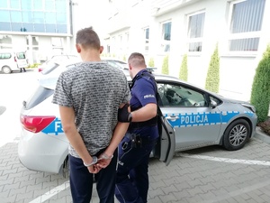 Umundurowany policjant  trzyma pod rekę skutego kajdankami mężczyznę. Dochodzą do radiowozu policyjnego.